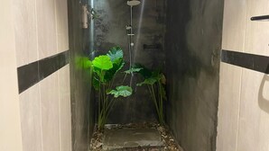 A narrow, enclosed space with a showerhead mounted on the ceiling, surrounded by large green plants.