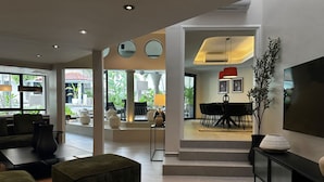 Een moderne woonkamer met een bank, voetenbankje en een televisie op een console. Er is een eethoek met een tafel en stoelen, en een trap die naar een ander niveau leidt.
