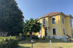 A yellow building with a red roof, a large tree in front, and a small garden area.