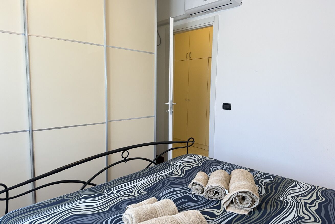 A bed with a patterned bedspread and rolled towels, a wardrobe with sliding doors, and a door leading to a room.