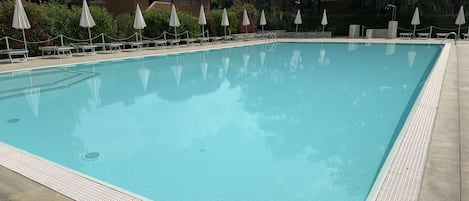 A rectangular swimming pool with a tiled edge, surrounded by lounge chairs and umbrellas, with a building in the background.