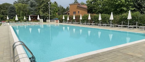 Una piscina rectangular con una solana empavedada, rodeada de sombrillas blancas y sillas de descanso, con árboles y un edificio al fondo.