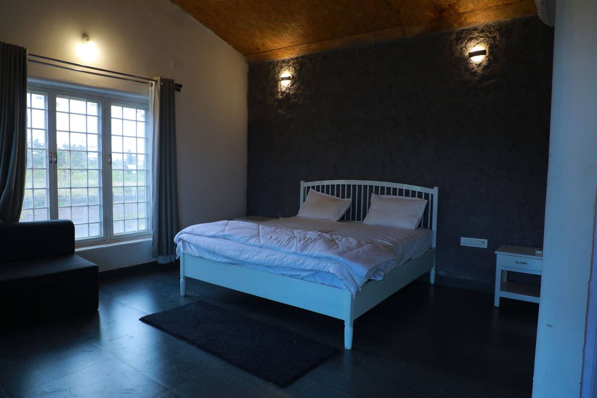 A bedroom with a bed, a nightstand, a window with curtains, and a view of the outdoors.