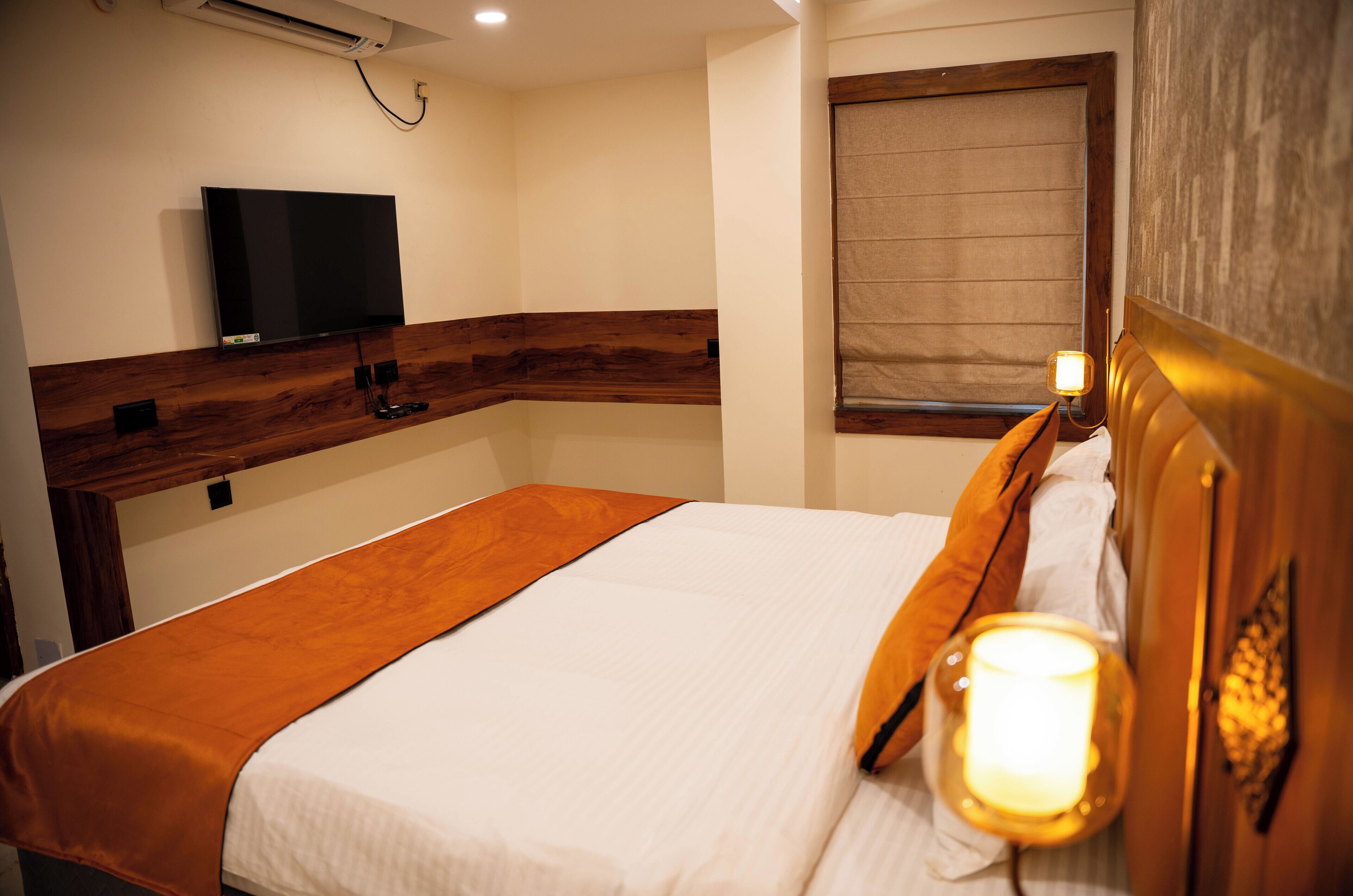 A hotel room with a bed, a television mounted on the wall, a window with a blind, and a bedside lamp.