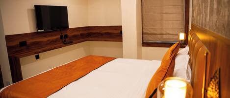 A hotel room with a bed, a television mounted on the wall, a window with a blind, and a bedside lamp.