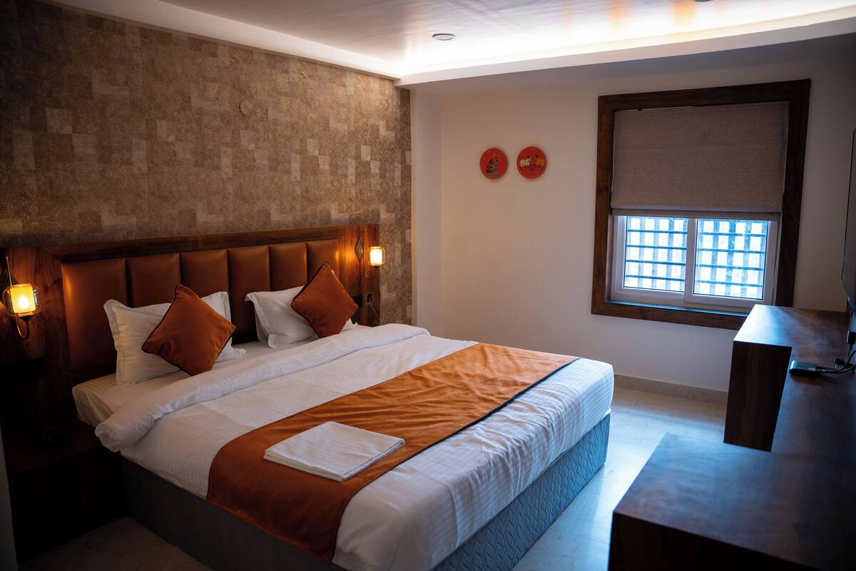 A neatly made bed with a headboard, a bedside table, and a window with blinds.