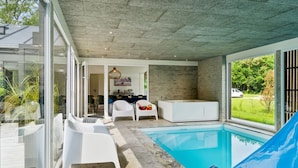 Un área de piscina interior moderna con techo de hormigón, mobiliario blanco y vistas al exterior.