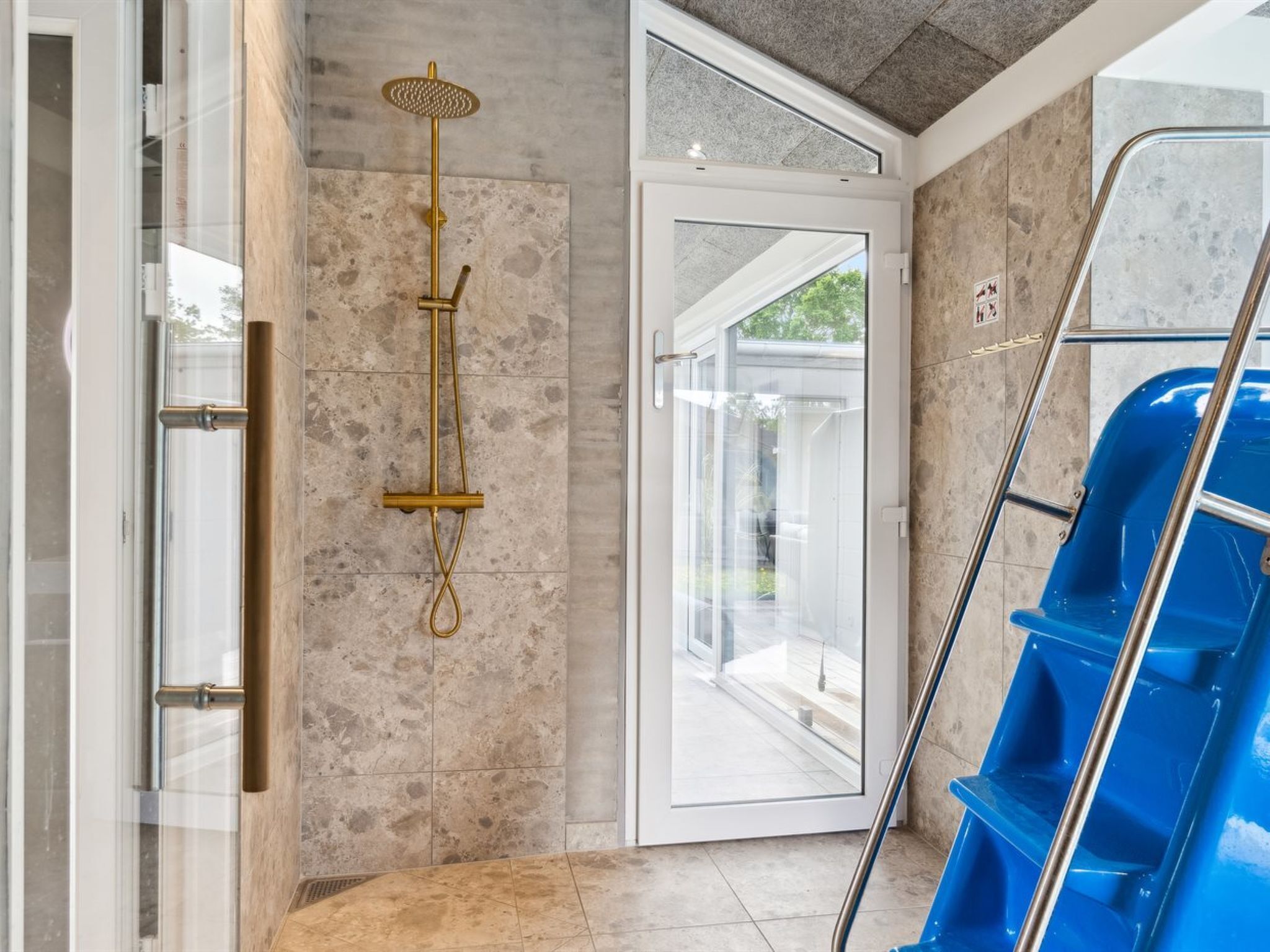 A modern bathroom with a glass door leading to an outdoor area, a gold showerhead, and a blue chair.