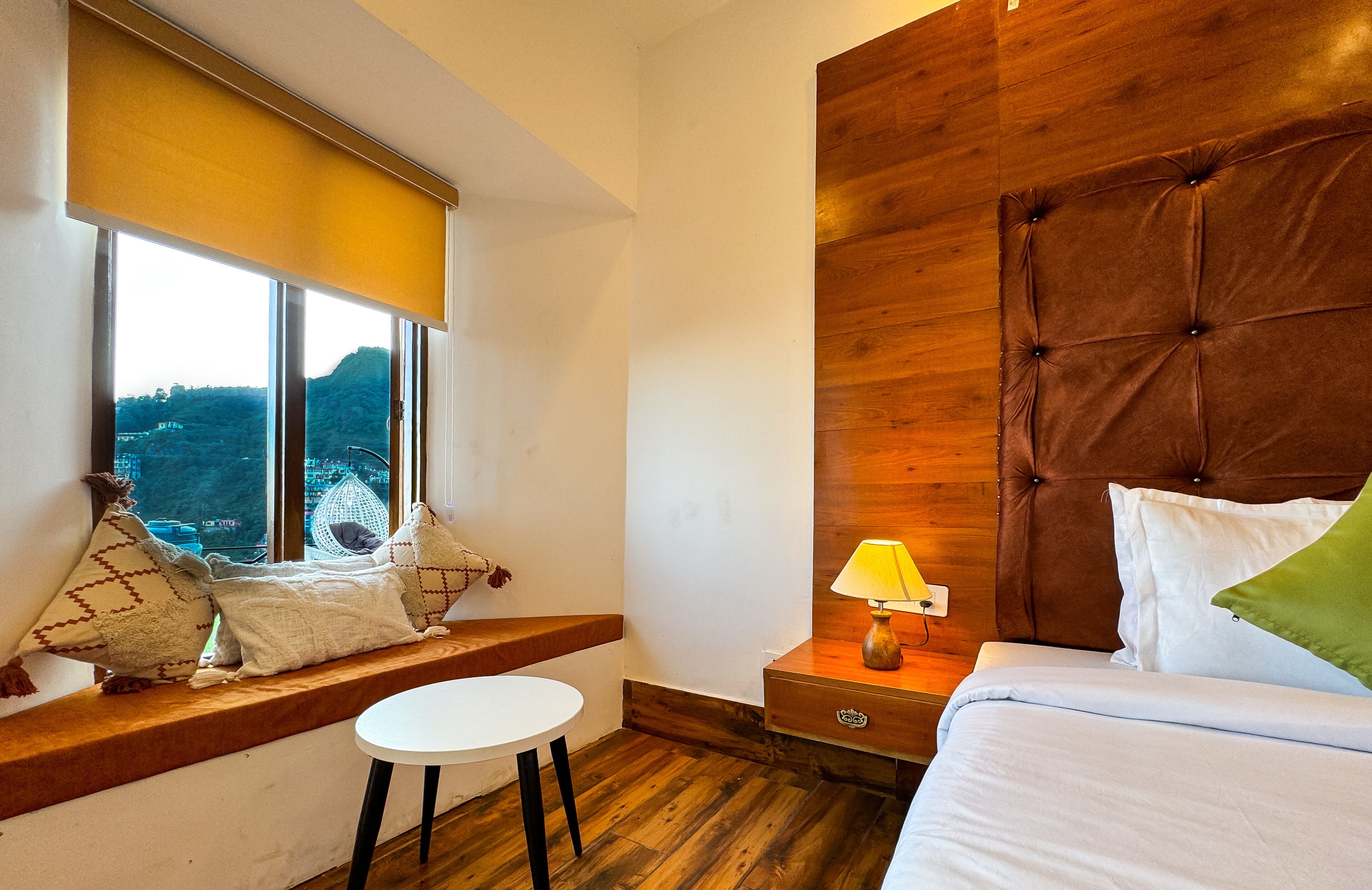 Une chambre avec une grande fenêtre, une tête de lit en bois, une table de chevet avec une lampe, et une vue sur l’extérieur.