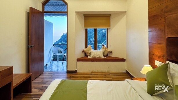 A bedroom with a wooden bed, a window seat with cushions, a wooden headboard, and a view of a mountainous landscape. - Hotel Wraveler Inn Mussorrie (Dehradun)