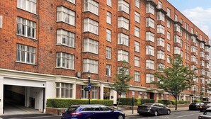 A multi-story brick building with a row of windows on the ground floor, a visible entrance, and a street view with parked cars.