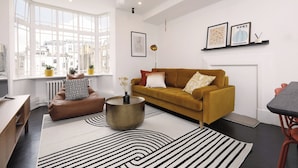 A modern living room with a mustard sofa, black and white striped rugs, and a large window offering a city view.