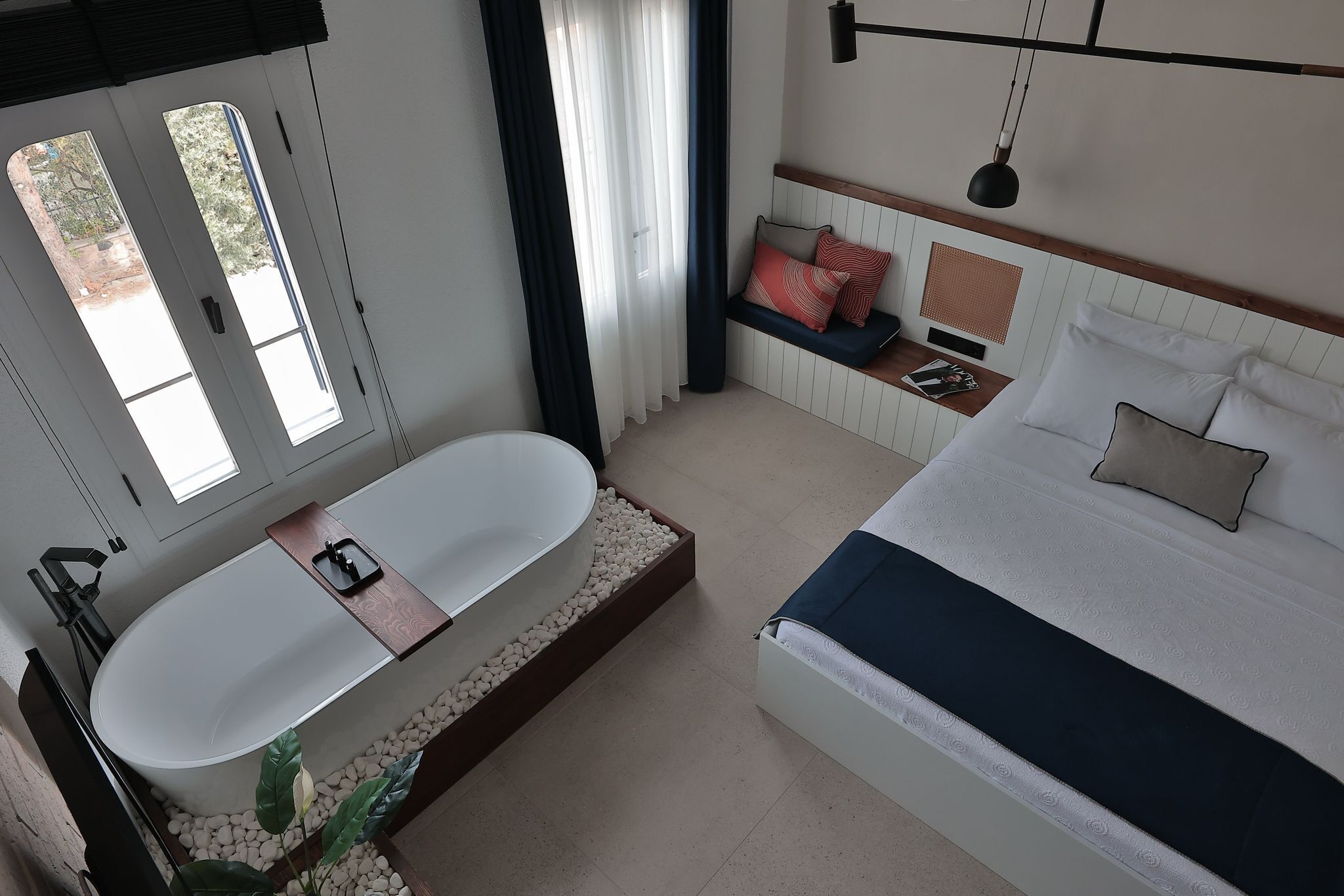 Une chambre à coucher moderne avec un grand lit, une banquette intégrée agrémentée de coussins, une baignoire blanche et une fenêtre avec des rideaux.