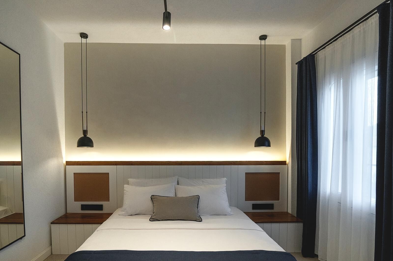 Una habitación de hotel moderna con una cama grande, dos mesitas de noche, cabecero con iluminación integrada y un espejo grande en una pared.