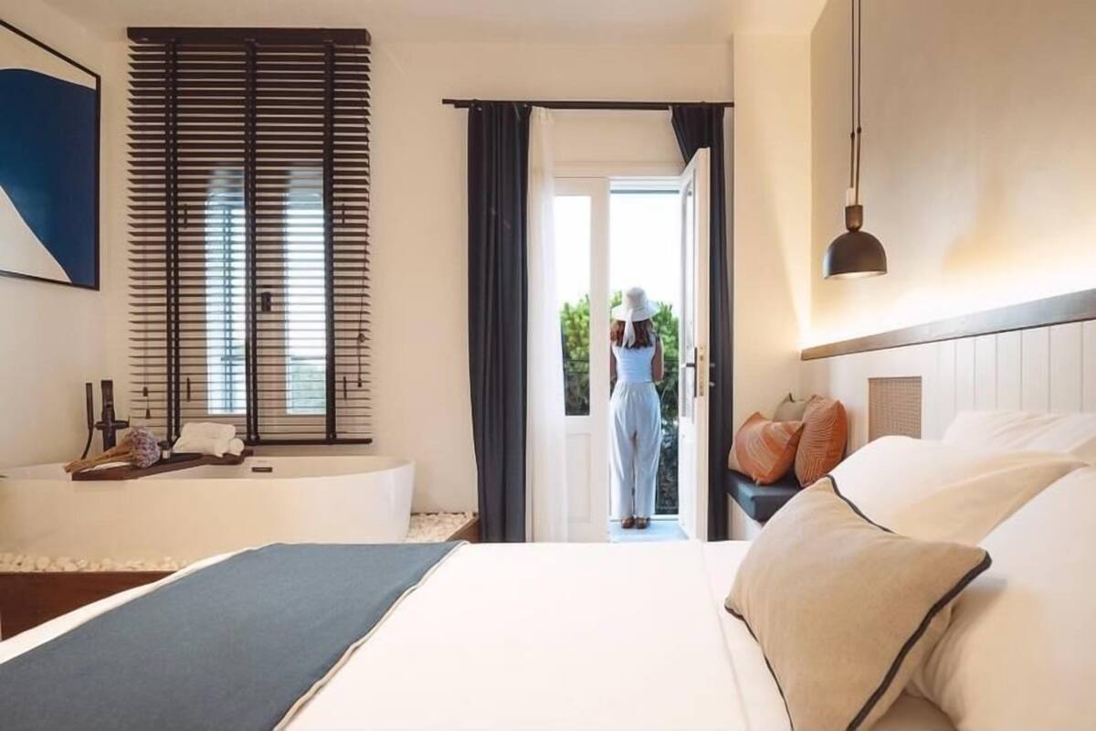 A hotel room with a bed, a bedside table, a window with blinds, a door leading outside, and a view of greenery outside.