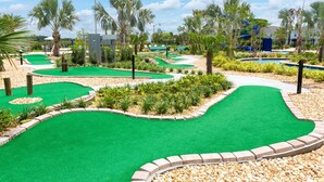 Un parcours de mini-golf doté de plusieurs zones d’entraînement au putting, entouré de palmiers et bordé d’une piscine.