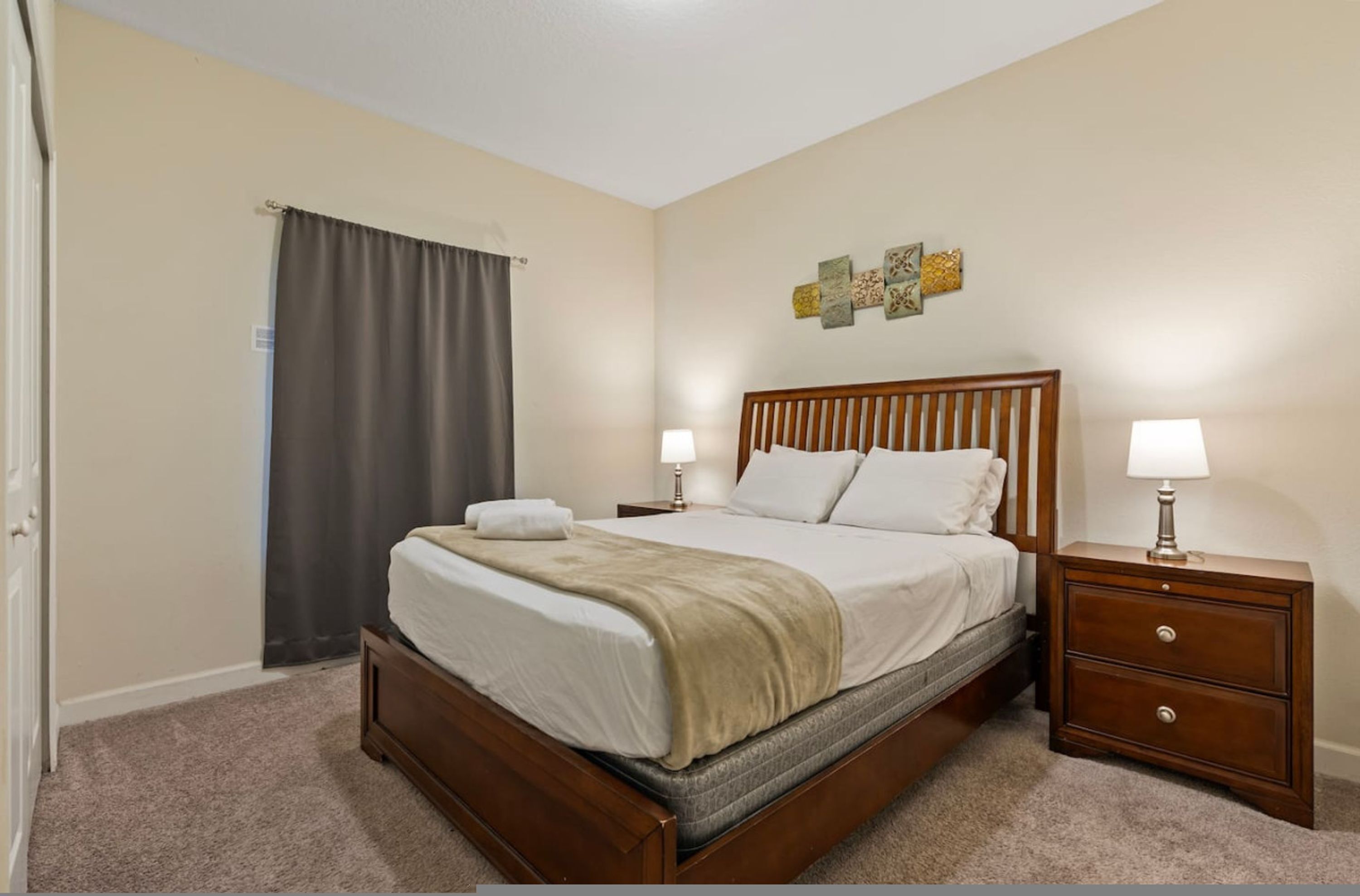A bedroom with a wooden bed, two bedside tables with lamps, a gray curtain, and a wall with decorative art.