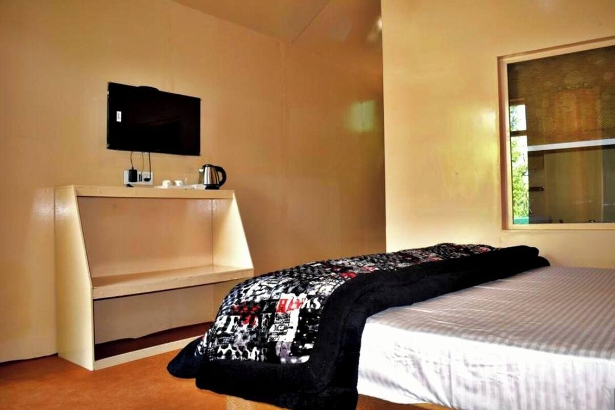 A bedroom with a bed, a TV mounted on the wall, a small table with a kettle, and a window.