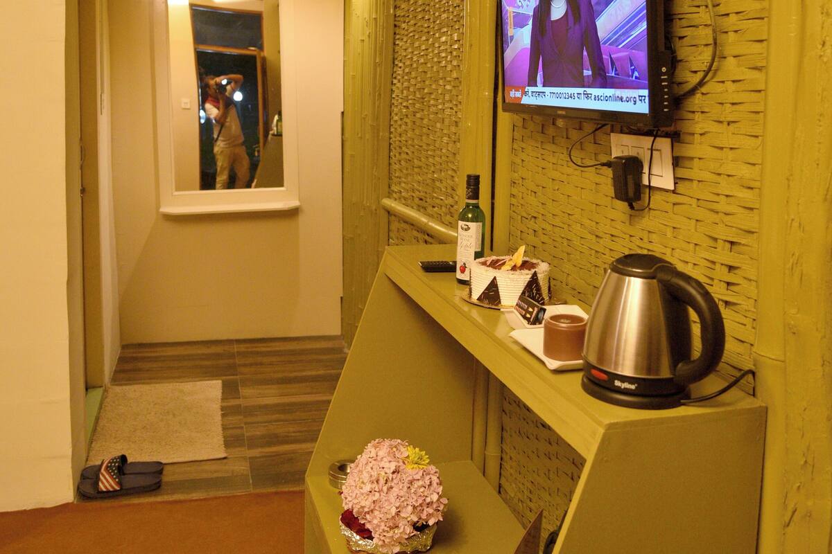 A reception area with a television displaying a program, a mirror, a small table with a bottle, a kettle, and a decorative item.