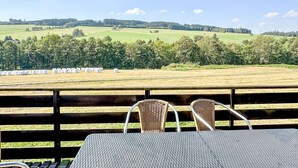 A view from a balcony with wicker chairs and a table, overlooking a green field with hay bales and trees.