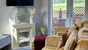 A living room with a sofa, a flat-screen TV, a fireplace, and a view of the outdoors. - Relax at our Lodge in the Cheshire Countryside (Congleton)