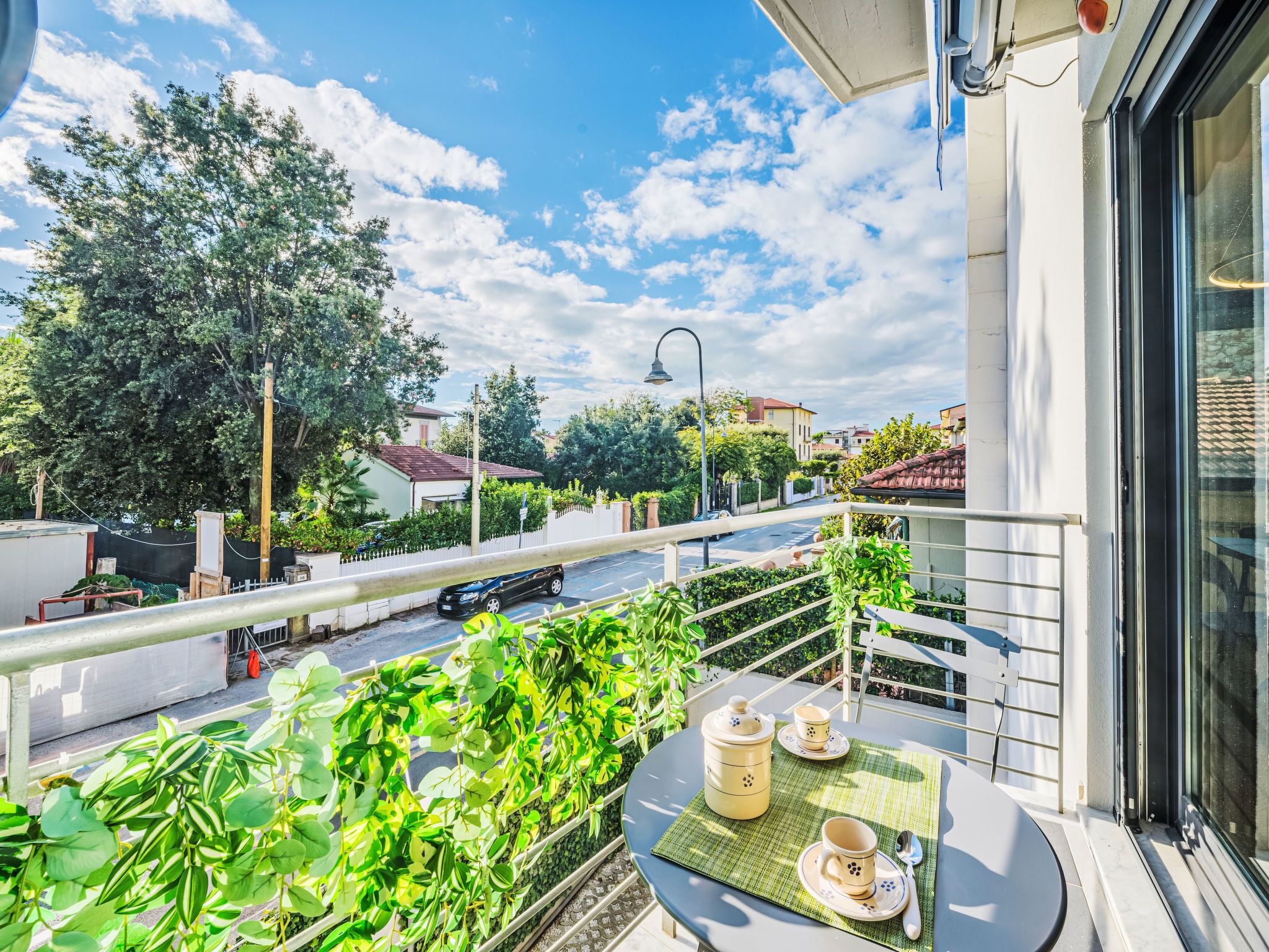 Ein Balkon mit einem für Tee gedeckten Tisch, mit Blick auf eine Straße mit Häusern und Grünpflanzen.