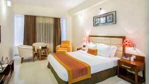 A hotel room with a bed, two chairs, a bedside table, a lamp, and a window with curtains. - Grand Harmony Beach Resort (Neyyattinkara)