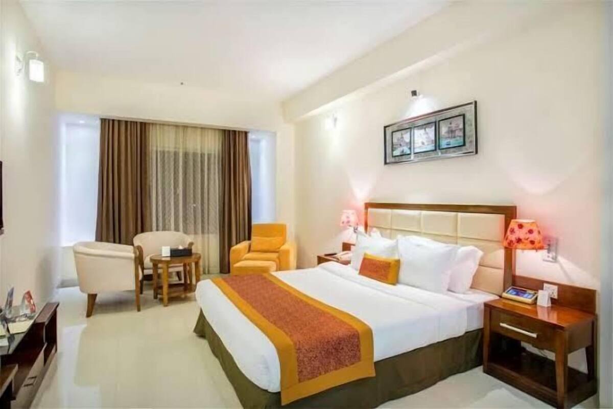 A hotel room with a bed, two chairs, a bedside table, a lamp, and a window with curtains.