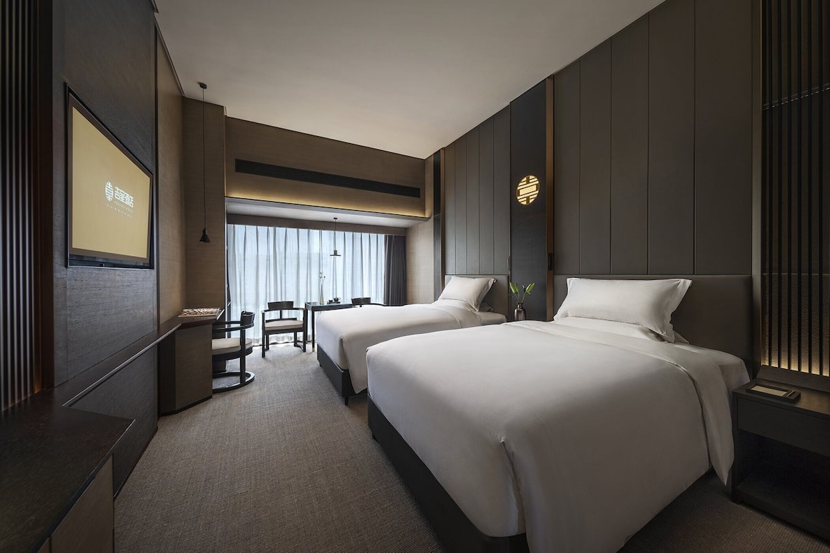 A modern hotel room with two beds, a desk, and a large window with curtains.