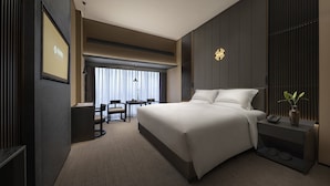 A modern hotel room with a large bed, a desk, and a chair. There is a window with curtains, a vase with a plant, and a wall-mounted light fixture.