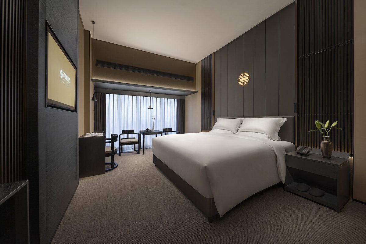 A modern hotel room with a large bed, a desk, and a chair. There is a window with curtains, a vase with a plant, and a wall-mounted light fixture.