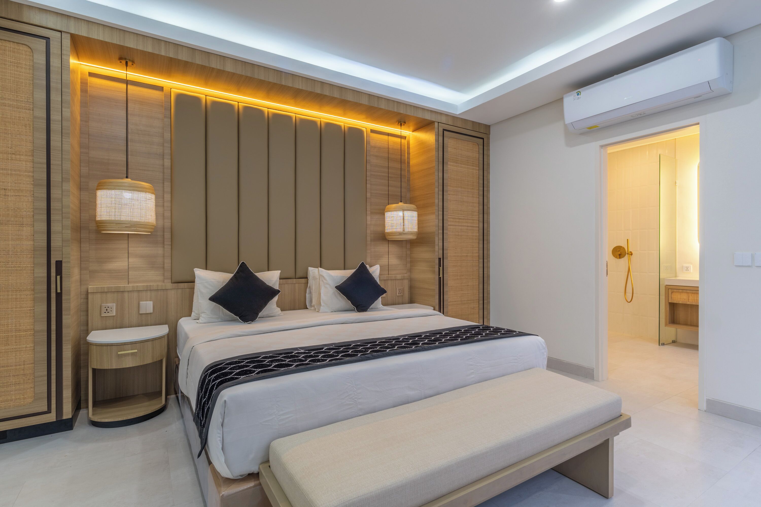 Quarto de hotel moderno com uma cama grande, abajures, mesa de cabeceira e um banco.