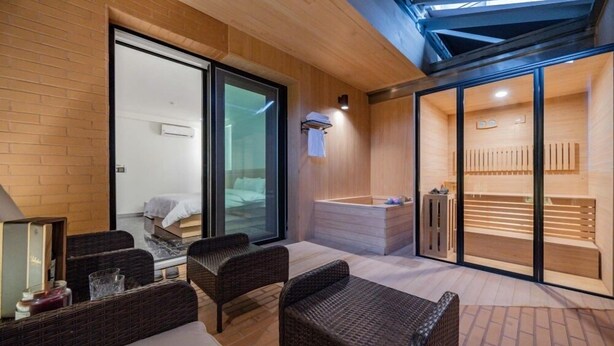 Habitación de hotel moderna con puerta de vidrio que da a un baño privado, una zona de estar con muebles de mimbre y un área de sauna visible.