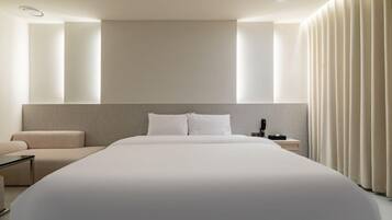 A modern hotel room with a large bed, a bedside table, a chair, and a large window with vertical blinds.