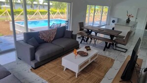 A modern living room with a gray sofa, a white coffee table, and a wooden dining table. There is a large window offering a view of a swimming pool and greenery. - Te Aro Villa 2
Modern Brand New 3brm villa with pool in Muri (Ngatangiia District)