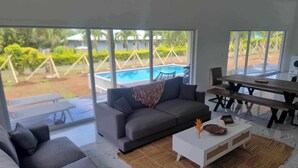 A modern living room with a grey sofa, a white coffee table, and a dining table with benches. Large windows offer a view of a swimming pool and greenery outside. - Te Aro Villa 2
Modern Brand New 3brm villa with pool in Muri (Ngatangiia District)