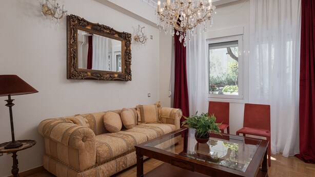 Un salon comprenant un canapé, une table basse avec un plateau en verre, des fauteuils rouges, un lustre, un grand miroir et une fenêtre avec des rideaux.