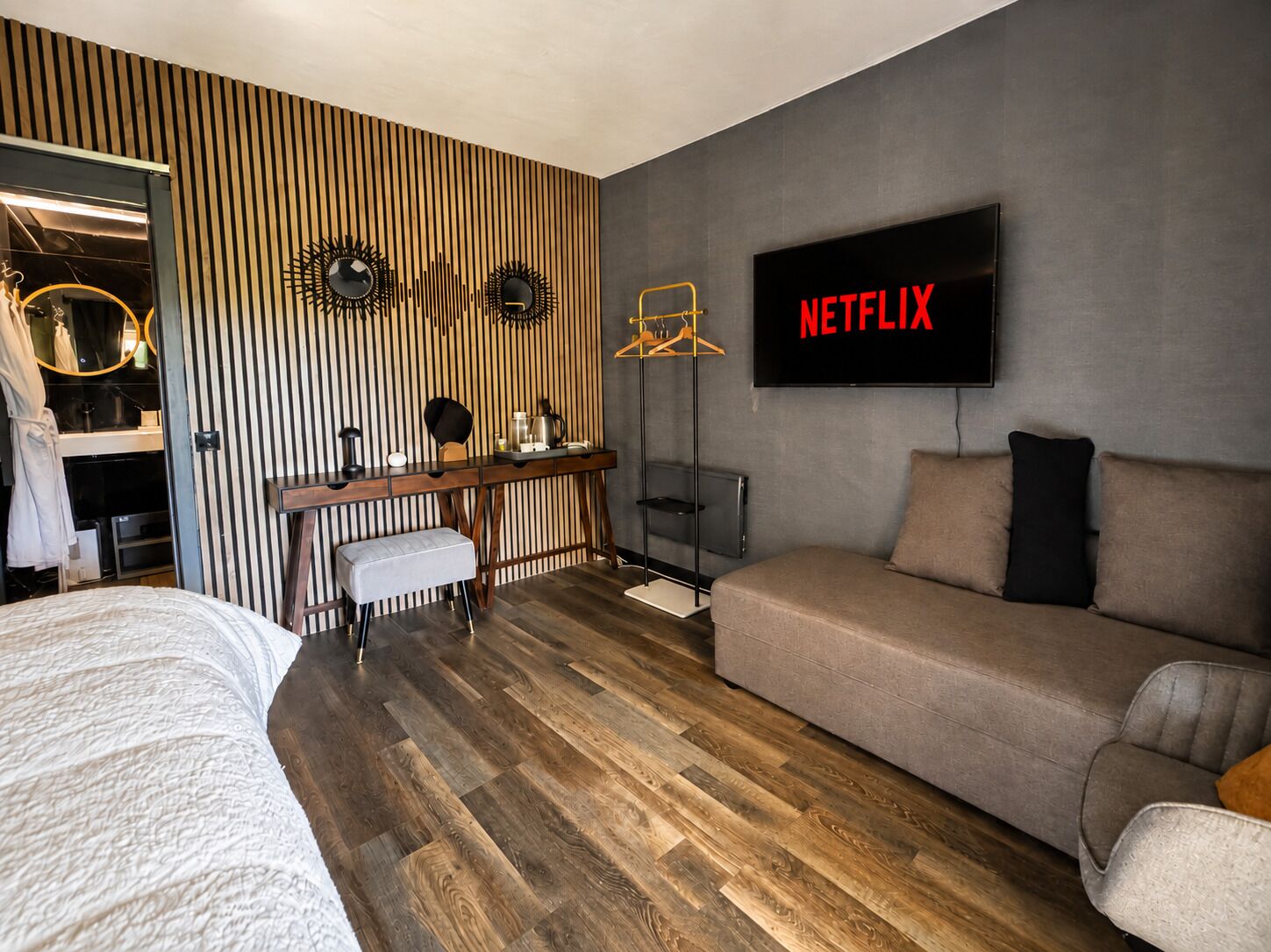 A modern bedroom with a wooden floor, a bed, a desk with a chair, a sofa, and a TV displaying 'NETFLIX'.