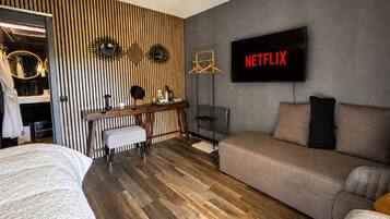 A modern bedroom with a wooden floor, a bed, a desk with a chair, a sofa, and a TV displaying 'NETFLIX'.