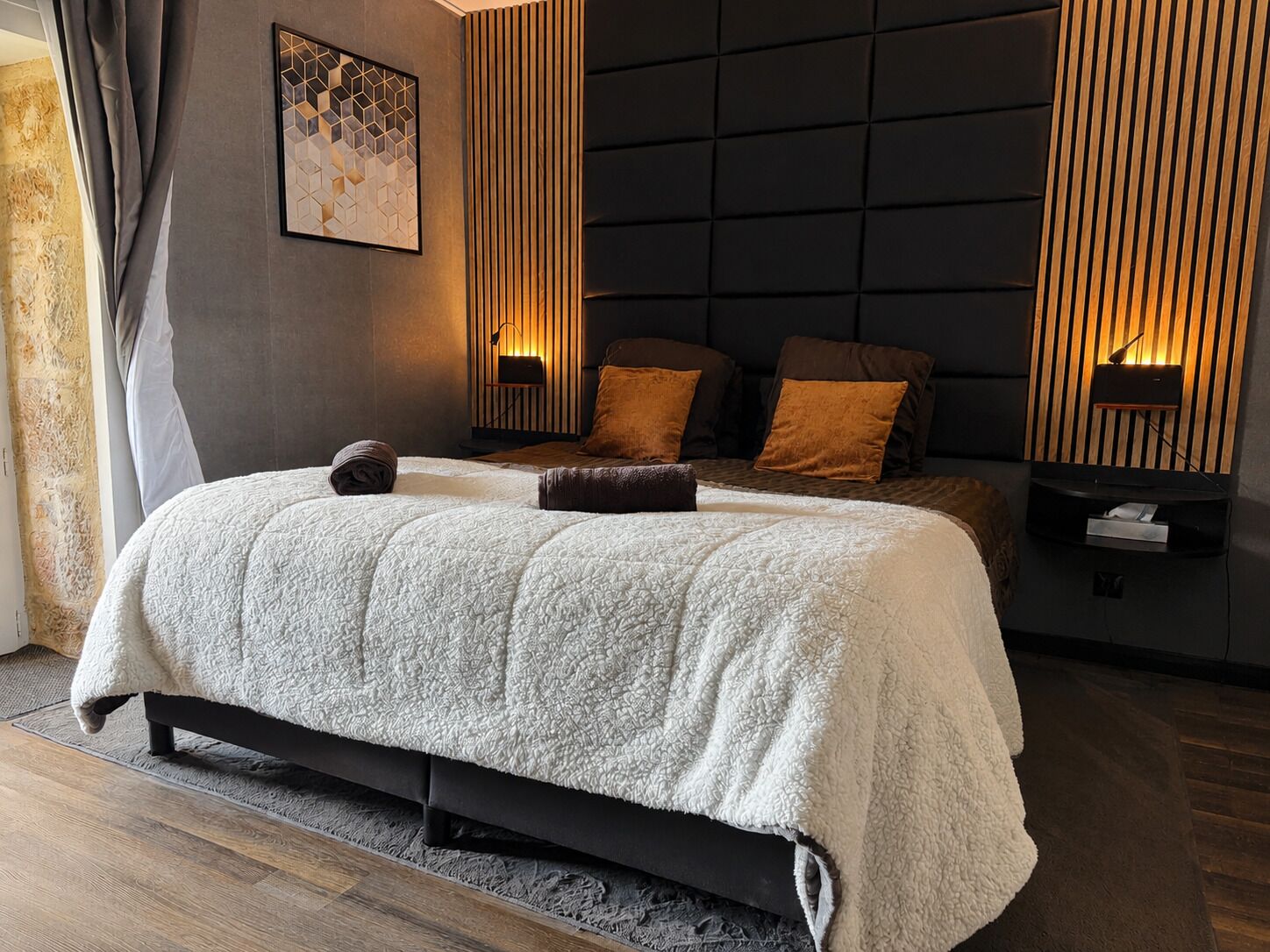 A modern bedroom with a large bed, a headboard with vertical slats, a wall-mounted light, and a framed geometric artwork.