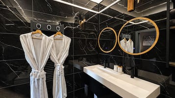 A modern bathroom with a black marble wall, two white robes hanging on a rack, a white sink, and two round mirrors.