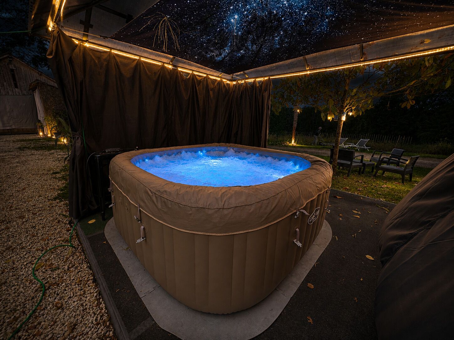 Un jacuzzi debajo de una carpa con luces colgantes, rodeado por un área de grava y una terraza de madera.