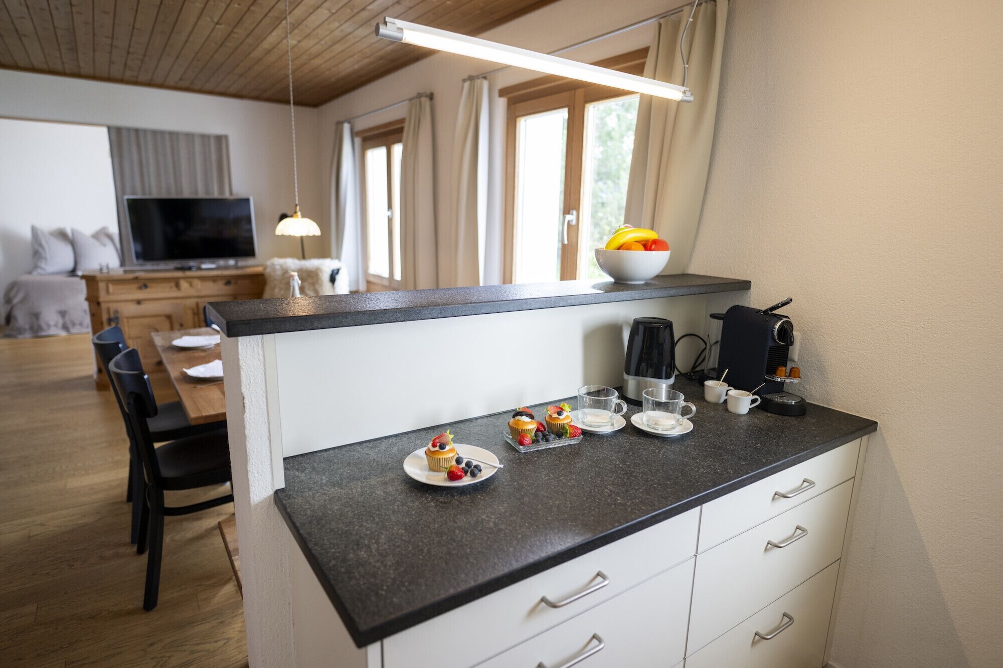 A compact kitchen with a dark countertop, white cabinets, and a coffee machine. There is a dining area with a table and chairs, and a TV in the background.
