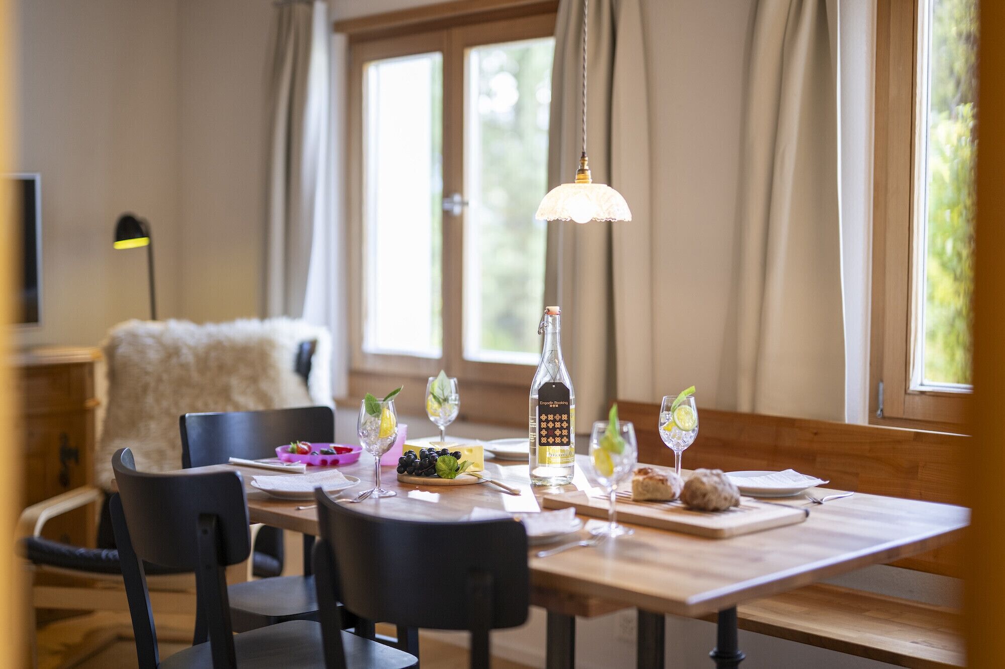 A dining area with a wooden table set for four, including wine glasses, a bottle of liquor, and a bowl of fruit.