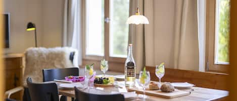 A dining area with a wooden table set for four, including wine glasses, a bottle of liquor, and a bowl of fruit.