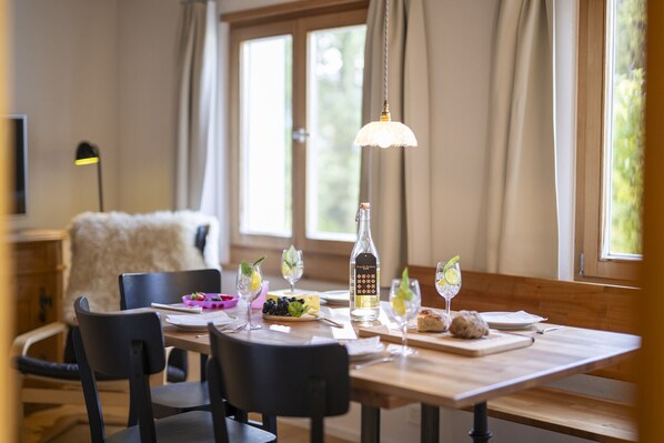 A dining area with a wooden table set for four, including wine glasses, a bottle of liquor, and a bowl of fruit.