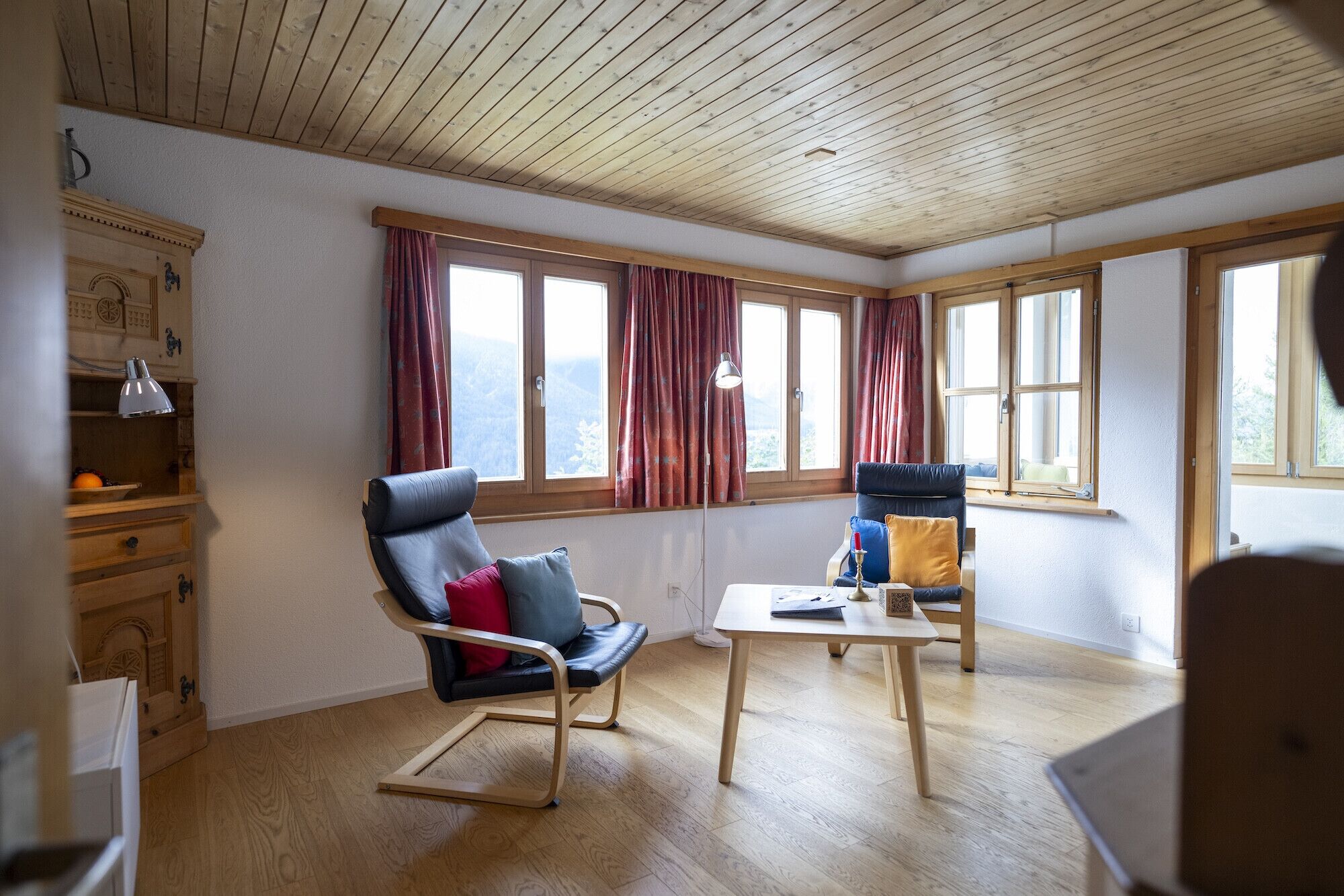 A room with a wooden ceiling, large windows with red curtains, a black leather chair, a wooden table, and a wooden cabinet with a lamp.