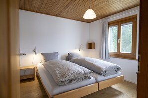 Un dormitorio con techo de madera, una cama con dos almohadas, una mesita de noche, una lámpara de pared y una ventana con cortinas.