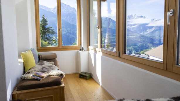 Une pièce chaleureuse avec un banc en bois, une petite table et une vue sur des montagnes enneigées, visible à travers de grandes fenêtres.