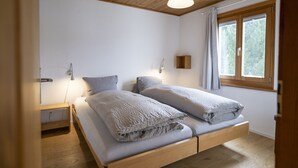 Une chambre avec un plafond en bois, un lit avec deux oreillers, une table de chevet, une lampe fixée au mur et une fenêtre avec des rideaux.